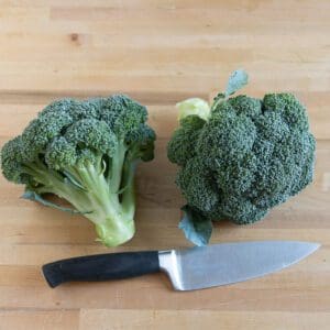 Two broccoli heads and a chef's knife on a wooden surface.
