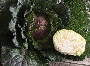 A purple cabbage with green leaves and a halved white cabbage on a wooden surface.