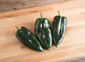 Three fresh green poblano peppers on a wooden surface.